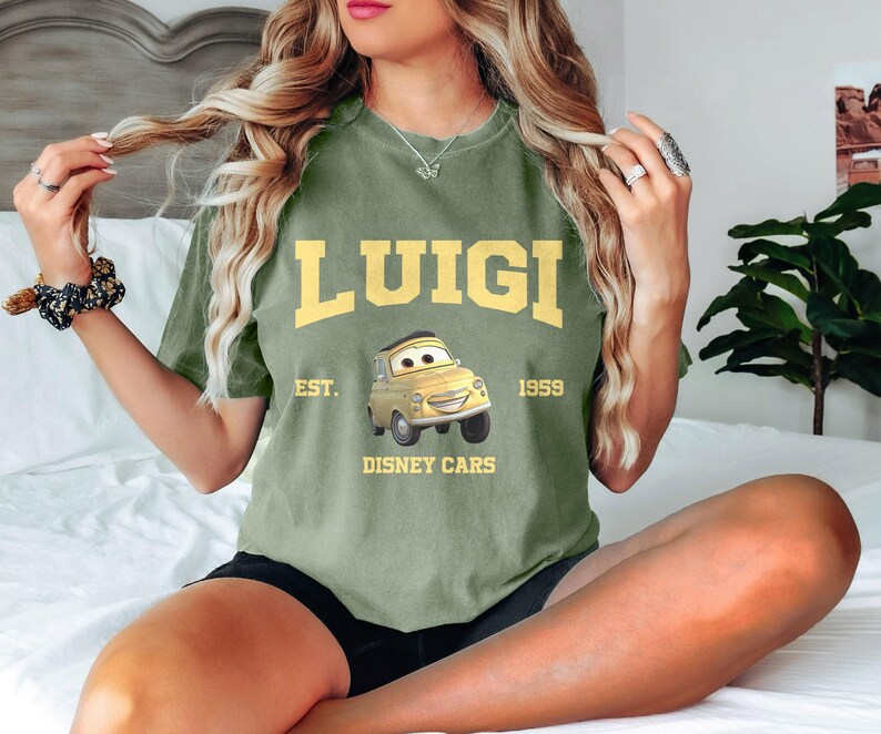 Puede incluir: Camiseta verde de manga corta con un coche de dibujos animados amarillo y el texto "LUIGI EST. 1959 DISNEY CARS".