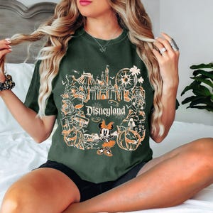 May include: Dark green t-shirt featuring a Disneyland graphic with orange and white illustrations of park icons, pumpkins, and Minnie Mouse. The word "Disneyland" is printed in white. The shirt is a relaxed fit.
