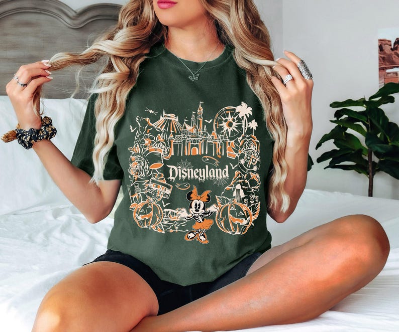May include: Dark green t-shirt featuring a Disneyland graphic with orange and white illustrations of park icons, pumpkins, and Minnie Mouse. The word "Disneyland" is printed in white. The shirt is a relaxed fit.