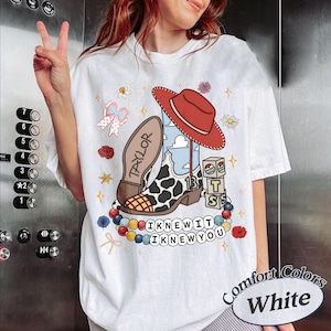 May include: A white t-shirt featuring a graphic print of a red cowboy hat, a cow-print boot with the number 13, blocks with the letters T, S, and the name TAYLOR. Below are colourful beads and the text I KNEW IT I KNEW YOU surrounded by flowers.
