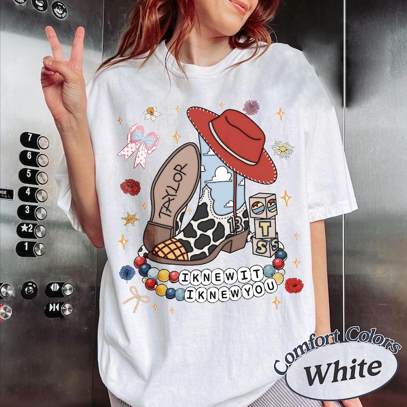 May include: A white t-shirt featuring a graphic print of a red cowboy hat, a cow-print boot with the number 13, blocks with the letters T, S, and the name TAYLOR. Below are colourful beads and the text I KNEW IT I KNEW YOU surrounded by flowers.