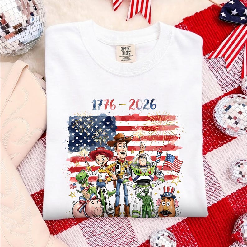 May include: A white t-shirt featuring a graphic of Toy Story characters including Woody, Buzz Lightyear, Jessie, Rex, Hamm, and a green soldier. The design includes a watercolor American flag, fireworks, and the text 1776 - 2026 in blue and red.
