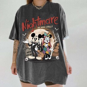 May include: Dark gray t-shirt featuring a graphic of Mickey and Minnie Mouse dressed as characters from The Nightmare Before Christmas. The shirt has the words "Nightmare on Main Street" in red, with a full moon and Halloween imagery.