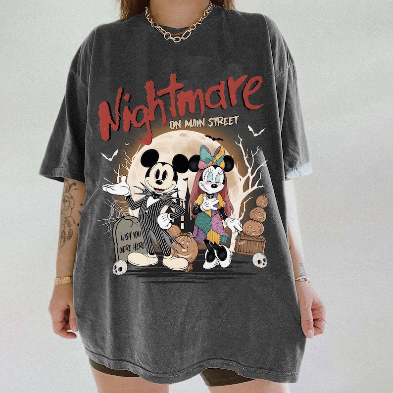 May include: Dark gray t-shirt featuring a graphic of Mickey and Minnie Mouse dressed as characters from The Nightmare Before Christmas. The shirt has the words "Nightmare on Main Street" in red, with a full moon and Halloween imagery.