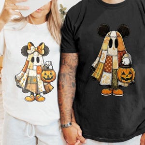以下が含まれることがあります： ミッキーマウスの耳を持つ漫画の幽霊のキャラクターが、パッチワークの衣装を着て、カボチャのランタンを持っている白と黒のTシャツ。幽霊はオレンジ色の靴とリボンをしています。黒いシャツも同じデザインです。