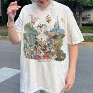 K&ouml;nnte beinhalten: Cremefarbenes T-Shirt mit einer farbenfrohen Grafik von Disney-Figuren, darunter Mickey Mouse, Minnie Mouse und Dumbo. Die Grafik enth&auml;lt auch den Text "Magic Kingdom Walt Disney World".