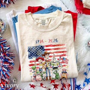 May include: A beige crew neck t-shirt featuring a graphic of Toy Story characters including Woody, Jessie, Buzz Lightyear, Rex, Hamm, and Mr. Potato Head in front of a watercolor American flag with fireworks and the text 1776 - 2026.