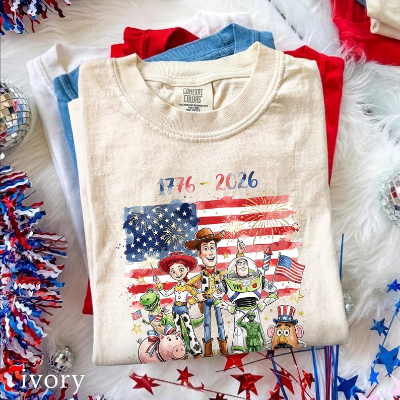 May include: A beige crew neck t-shirt featuring a graphic of Toy Story characters including Woody, Jessie, Buzz Lightyear, Rex, Hamm, and Mr. Potato Head in front of a watercolor American flag with fireworks and the text 1776 - 2026.