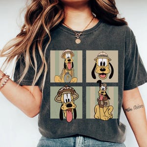 May include: Dark grey t-shirt with a four-panel design of Pluto in safari gear. Each panel shows Pluto in different poses, wearing a safari hat and backpack. The design is set against a striped background.