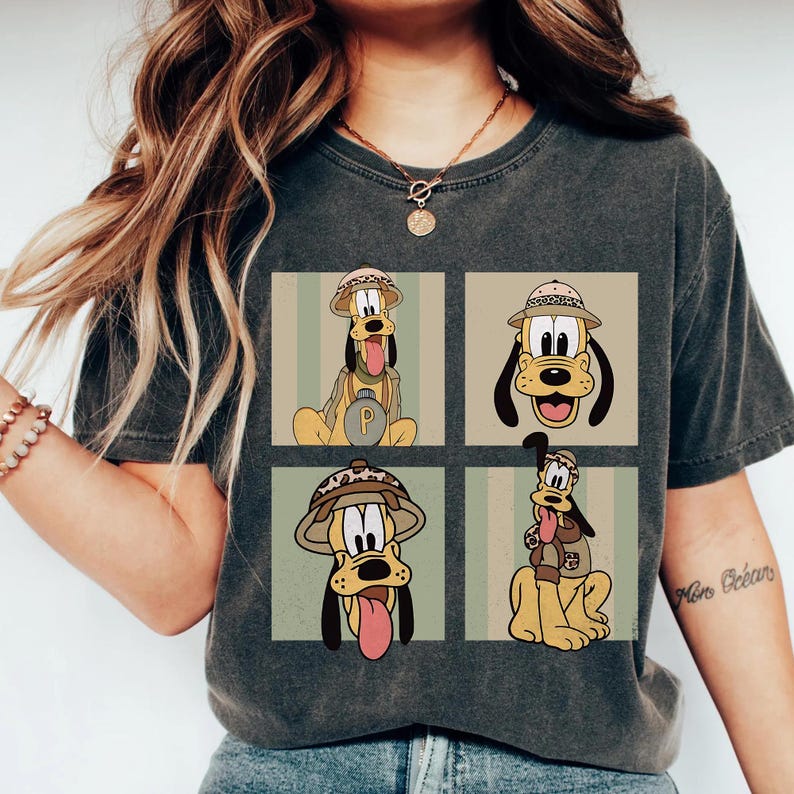 May include: Dark grey t-shirt with a four-panel design of Pluto in safari gear. Each panel shows Pluto in different poses, wearing a safari hat and backpack. The design is set against a striped background.