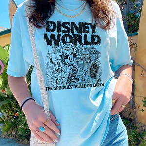 K&ouml;nnte beinhalten: Ein hellblaues T-Shirt mit einem schwarzen Grafikdruck von Mickey Mouse und Minnie Mouse vor einer gruseligen Version von Disney World. Der Text "Disney World" befindet sich &uuml;ber den Figuren und "The Spookiest Place on Earth" darunter.