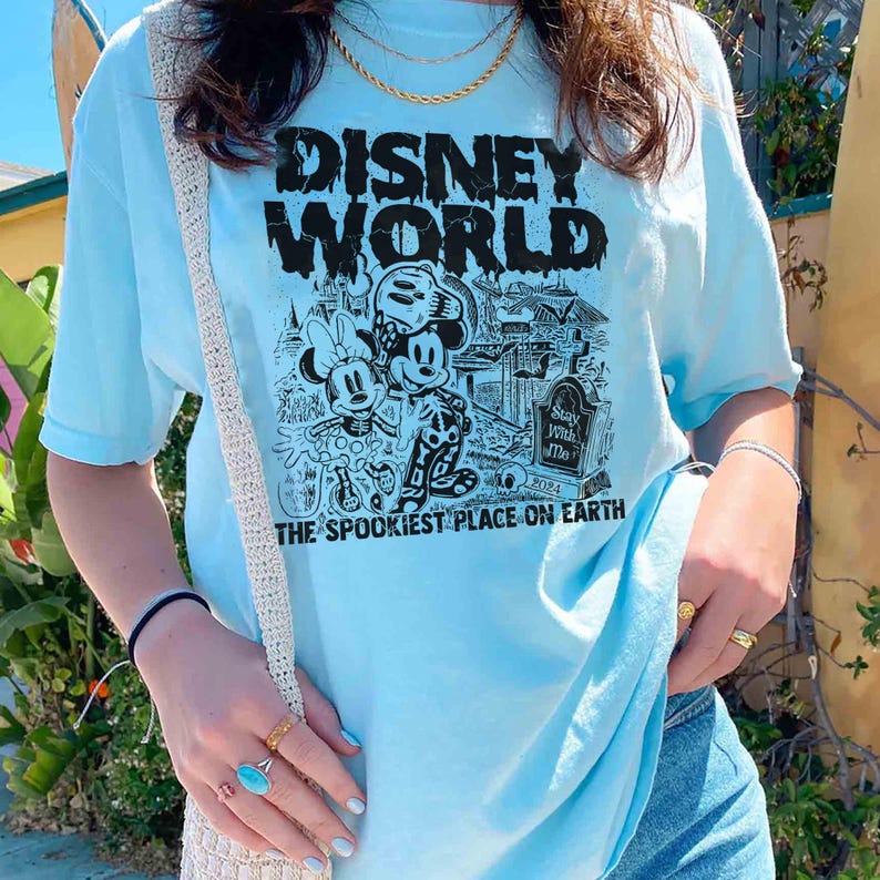 K&ouml;nnte beinhalten: Ein hellblaues T-Shirt mit einem schwarzen Grafikdruck von Mickey Mouse und Minnie Mouse vor einer gruseligen Version von Disney World. Der Text "Disney World" befindet sich &uuml;ber den Figuren und "The Spookiest Place on Earth" darunter.