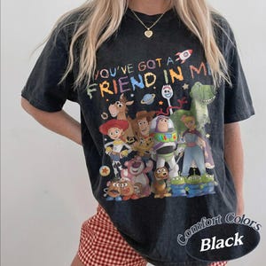 May include: A black mineral-washed t-shirt featuring a colorful graphic print of various animated characters and the text "YOU'VE GOT A FRIEND IN ME" at the top. The design includes a rocket, a planet, and multiple figures from a popular film franchise.