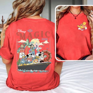 May include: Red t-shirt with a Disney cruise design. The back of the shirt shows the words "Disney Magic 2026" above a graphic of Disney characters on a ship. The front has a small graphic of Winnie the Pooh on a float.