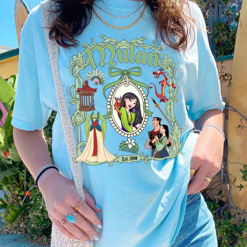 Puede incluir: Una camiseta azul claro con un estampado gr&aacute;fico de Mulan de la pel&iacute;cula de Disney Mulan. El gr&aacute;fico incluye a Mulan, Mushu, el drag&oacute;n y otros personajes de la pel&iacute;cula. El texto "Mulan" y "Est. 1998" tambi&eacute;n est&aacute;n incluidos en el gr&aacute;fico.