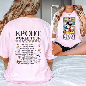 Pode incluir: Camiseta rosa com um gr&aacute;fico de Mickey Mouse e outros personagens da Disney. O texto "EPCOT World Tour" est&aacute; impresso na camiseta. A parte de tr&aacute;s da camiseta tem uma lista de pa&iacute;ses e cidades com o texto "EPCOT World Tour" impresso acima da lista.