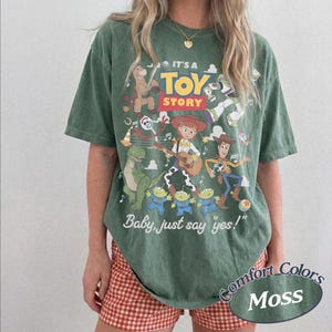 May include: A moss green short-sleeve t-shirt featuring a graphic print of Toy Story characters including Woody, Jessie, Rex, and Forky. The text reads IT'S A TOY STORY and Baby, just say yes! The shirt has a crew neckline and a relaxed, loose fit.