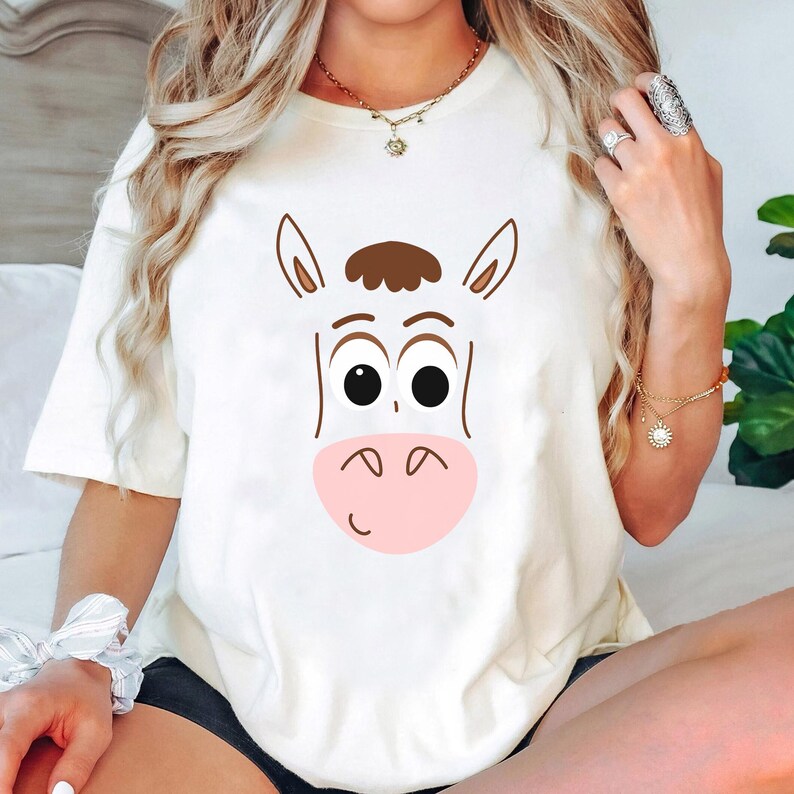 May include: Cream-colored t-shirt featuring a cartoon horse face with brown and black details. The horse has large black eyes, a pink muzzle, and a brown mane. The shirt is a casual, short-sleeved style.