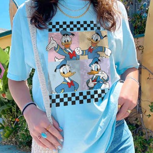 May include: Light blue t-shirt featuring a graphic of Donald Duck in four different poses. The graphic is set against a backdrop of pastel colors and black and white checkered patterns. The shirt has short sleeves and a crew neck.