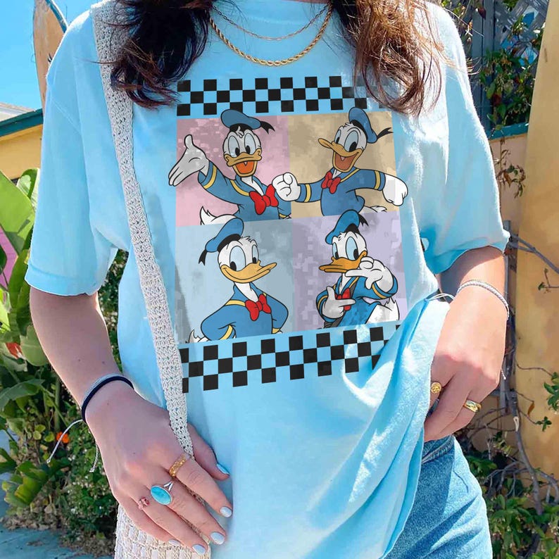 May include: Light blue t-shirt featuring a graphic of Donald Duck in four different poses. The graphic is set against a backdrop of pastel colors and black and white checkered patterns. The shirt has short sleeves and a crew neck.