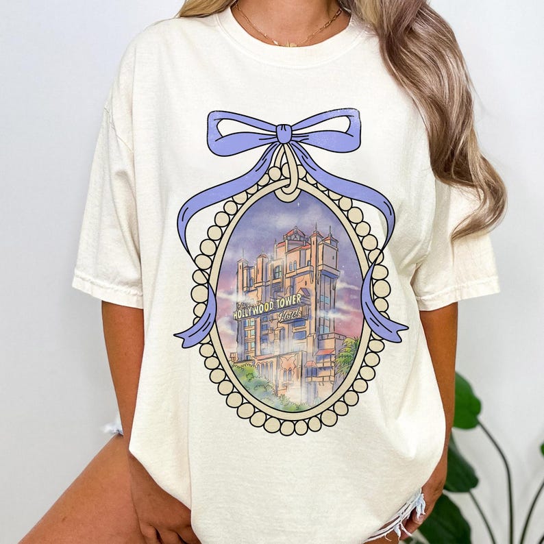 May include: A white t-shirt with a purple bow and a framed illustration of the Hollywood Tower Hotel. The illustration is in a vintage style and features a purple sky with clouds. The text "Hollywood Tower Hotel" is visible on the building.