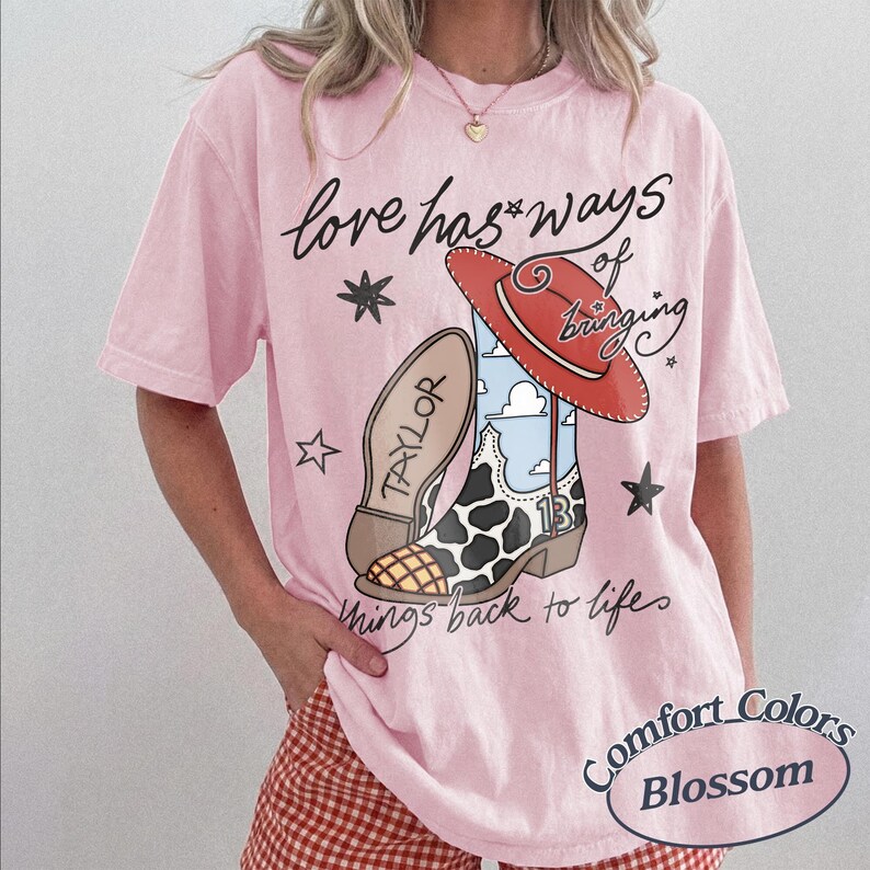 May include: A light pink t-shirt featuring a graphic of a cowboy boot with a cow print pattern, a red hat, and the text "love has ways of bringing things back to life" and "TAYLOR" on the boot sole. The boot also displays the number 13.