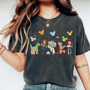 Puede incluir: Camiseta gris oscuro con un gr&aacute;fico colorido de personajes de dibujos animados, incluyendo un dinosaurio, un vaquero, un astronauta y un caballo. El dise&ntilde;o incluye globos con forma de Mickey Mouse y estrellas. La camiseta es de material suave.