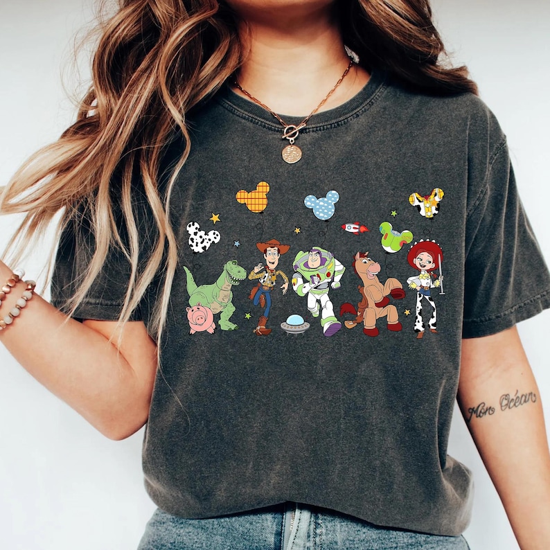 Puede incluir: Camiseta gris oscuro con un gr&aacute;fico colorido de personajes de dibujos animados, incluyendo un dinosaurio, un vaquero, un astronauta y un caballo. El dise&ntilde;o incluye globos con forma de Mickey Mouse y estrellas. La camiseta es de material suave.