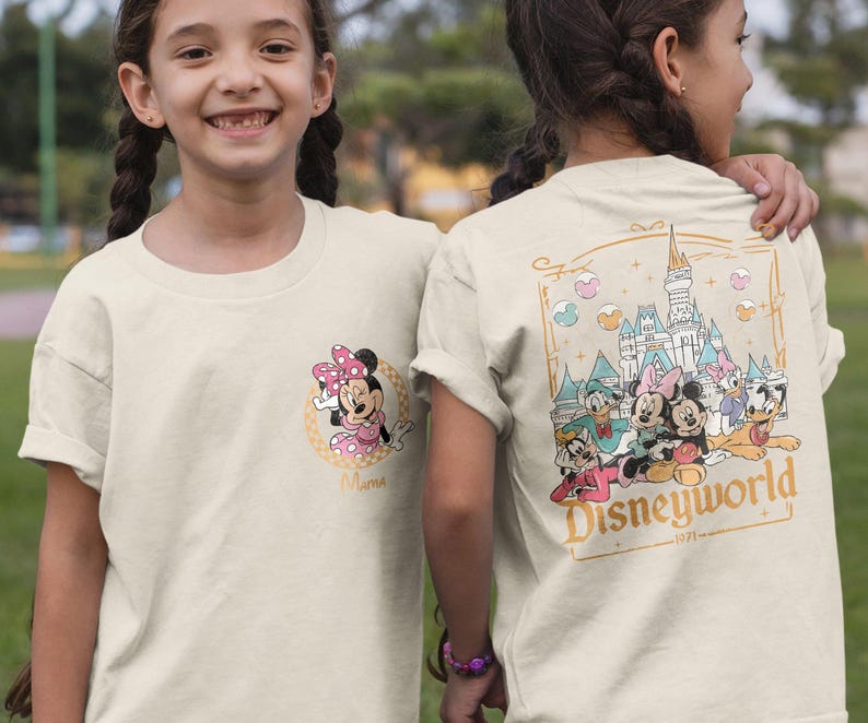 May include: Two children wearing cream colored t-shirts with a Disney World theme. One shirt features Mickey Mouse and Minnie Mouse, the other shirt features Mickey Mouse, Minnie Mouse, Donald Duck, Goofy, Pluto, and a castle with the text "Disneyworld 1971 - ".