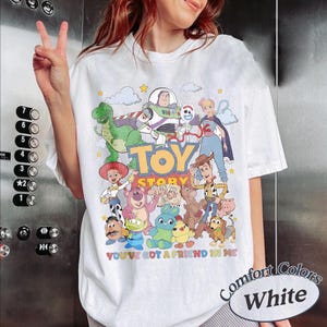 May include: A white t-shirt featuring a colorful graphic print of various Toy Story characters including Buzz Lightyear, Woody, Jessie, Rex, and others. The text "TOY STORY" is printed in large yellow letters, with "YOU'VE GOT A FRIEND IN ME" below.