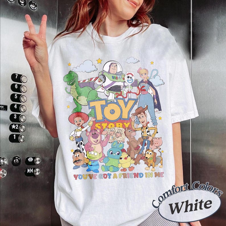 May include: A white t-shirt featuring a colorful graphic print of various Toy Story characters including Buzz Lightyear, Woody, Jessie, Rex, and others. The text "TOY STORY" is printed in large yellow letters, with "YOU'VE GOT A FRIEND IN ME" below.