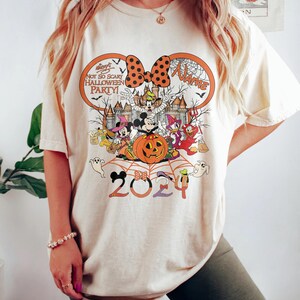 May include: Cream-coloured t-shirt with a Halloween design. The graphic includes Mickey Mouse, Minnie Mouse, Donald Duck, and other Disney characters. The text reads "Not So Scary Halloween Party!" and "2024". A large orange bow is at the top.