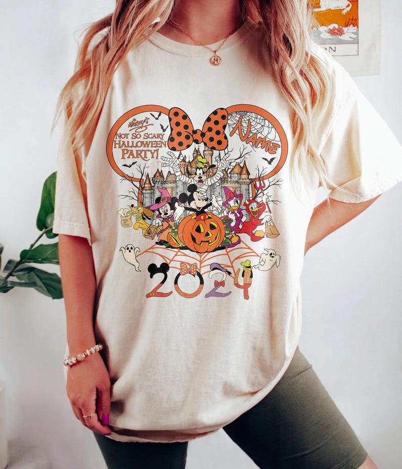 May include: Cream-coloured t-shirt with a Halloween design. The graphic includes Mickey Mouse, Minnie Mouse, Donald Duck, and other Disney characters. The text reads "Not So Scary Halloween Party!" and "2024". A large orange bow is at the top.