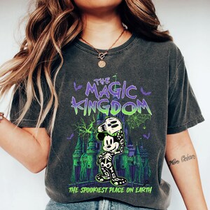 May include: Dark grey t-shirt with a Halloween graphic. The design features the text "THE MAGIC KINGDOM" in purple and green, a spooky Mickey Mouse character, and a castle. The phrase "THE SPOOKIEST PLACE ON EARTH" is also included.