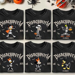 Op de afbeelding: Zes zwarte t-shirts met Halloween-thema Disney-personage-ontwerpen. Elk shirt heeft een ander personage in skeletvorm, met de tekst "Disneyworld" en "Florida 1971". Decoratieve pompoenen, spinnen en vleermuizen omringen de shirts.
