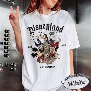 May include: A white t-shirt featuring a graphic print with the text Disneyland Est 1955 California. The design includes illustrations of Mickey Mouse, Minnie Mouse, Goofy, and Donald Duck dressed in Halloween costumes, including a mummy wrap.