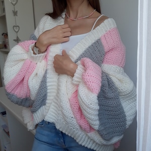 Könnte beinhalten: Ein handgefertigter, übergroßer Häkelcardigan in Weiß, Rosa und Grau. Der Cardigan mit offener Front hat weite Ärmel und ein strukturiertes Stichmuster. Das farbblockierte Design sorgt für einen gemütlichen und stilvollen Look. Dieses Kleidungsstück ist perfekt zum Überziehen.