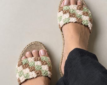 May include: A pair of slip-on sandals featuring a crochet upper with a checkered pattern in shades of sage green, cream, and brown. The footbed is lined with a woven tan material, and the edges are finished with a braided jute trim detail.