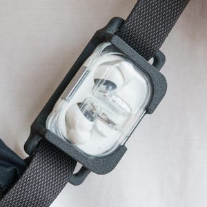 May include: A black and clear earbud case attached to a black strap, secured to a black bag. The case holds white earbuds. The strap is attached to a white shirt. A black bracelet is visible on the wrist.