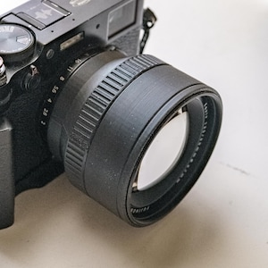 May include: A black Fujifilm X100V digital camera with a lens attached. The camera has several dials and buttons, including a red shutter button. The lens has a black lens hood. The camera is on a white surface.