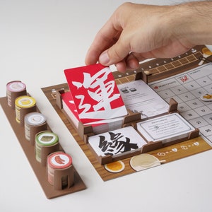 Puede incluir: Primer plano de un juego de mesa con un marco de madera y una cuadrícula de calendario. El juego incluye cartas rojas y negras con caracteres japoneses, y un estante de madera con fichas de colores. El juego se llama "168".