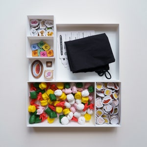 Puede incluir: Una caja blanca con varios compartimentos que contienen diferentes piezas de juego. Los compartimentos contienen pequeñas fichas de colores, una bolsa de tela negra, pequeños alimentos de plástico y un plato con un diseño rojo y blanco.