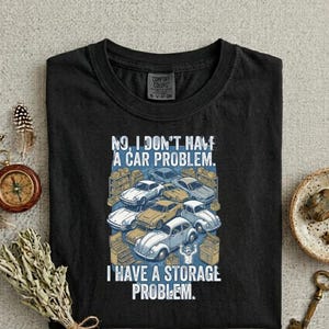 May include: A black t-shirt featuring a graphic print of various vintage cars parked in a garage setting. The text reads, "NO, I DON'T HAVE A CAR PROBLEM. I HAVE A STORAGE PROBLEM." The design includes multiple car illustrations in shades of blue, yellow, and white.