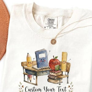Peut inclure: Un t-shirt couleur crème avec une étiquette Comfort Colors au col et un collier doré. Le motif présente une illustration aquarelle d'un bureau avec un crayon souriant, un cahier bleu, une pomme rouge à lunettes et une règle, avec le texte Custom Your Text.