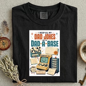 Peut inclure: Un t-shirt noir avec une impression graphique portant le texte 'I KEEP ALL MY DAD JOKES IN A DAD-A-BASE'. L'illustration comprend un ordinateur vintage, un magnétophone à bobines, une tasse de café et diverses icônes et étiquettes.
