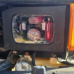 May include: Close-up of a vehicle's headlight assembly. The headlight is rectangular with a clear lens and a black surround. An amber turn signal is visible to the right. The vehicle's front end is partially visible.