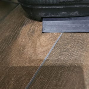 May include: A close-up of a black rubber doorstop with a rectangular shape. The doorstop is on a wood floor.