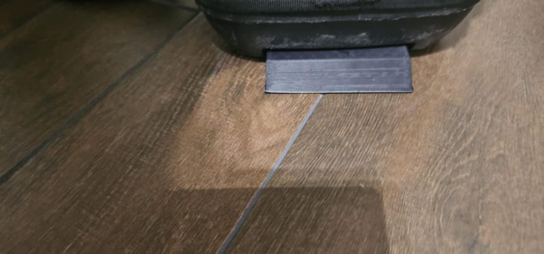 May include: A close-up of a black rubber doorstop with a rectangular shape. The doorstop is on a wood floor.