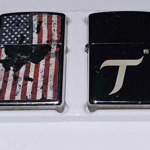 May include: Two silver lighters, one with a distressed American flag design and the other with a black background and a silver "T" logo.