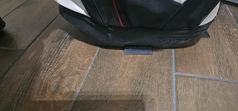 May include: A black and white golf bag with a red stripe and a black base. The bag is sitting on a wooden floor.